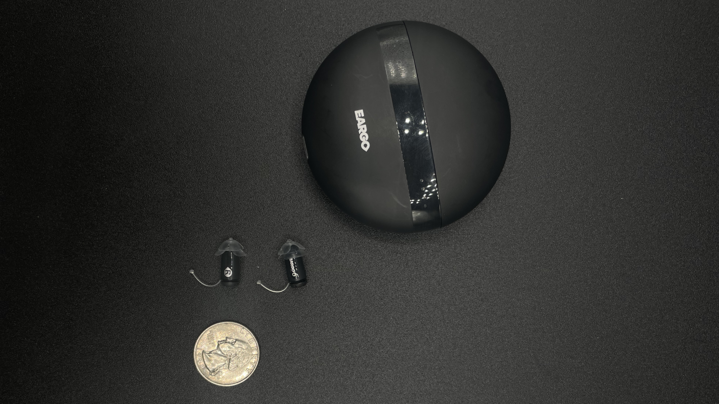 Eargo hearing aids and case next to American quarter coin for size; one Eargo hearing aid is about the size of a quarter
